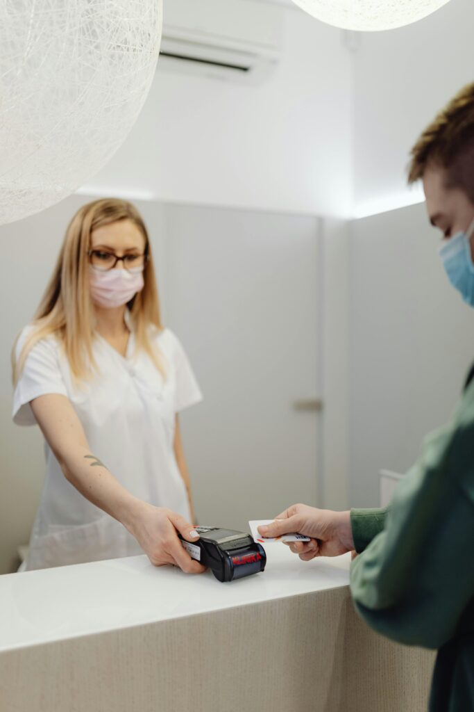 zipwp-image-6627907 A masked patient pays at a clinic counter, using a card terminal handled by a healthcare professional.