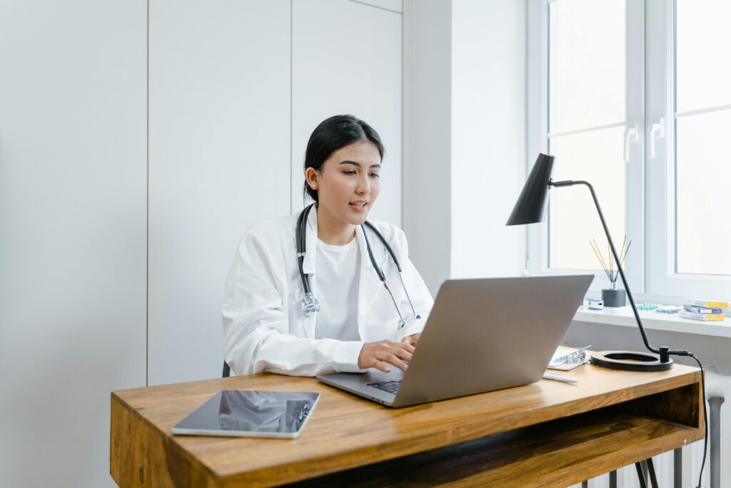 zipwp-image-8376232 A female doctor in a white coat uses a laptop for an online consultation from her office.