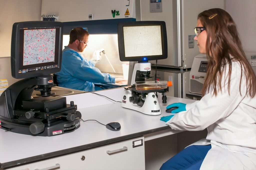 zipwp-image-s6w5noxtm7u A technician viewing a blot on a fluorescence microscope while another technician is using a pipette at the Advanced Technology Research Facility (ATRF), Frederick National Laboratory for Cancer Research, National Cancer Institute.